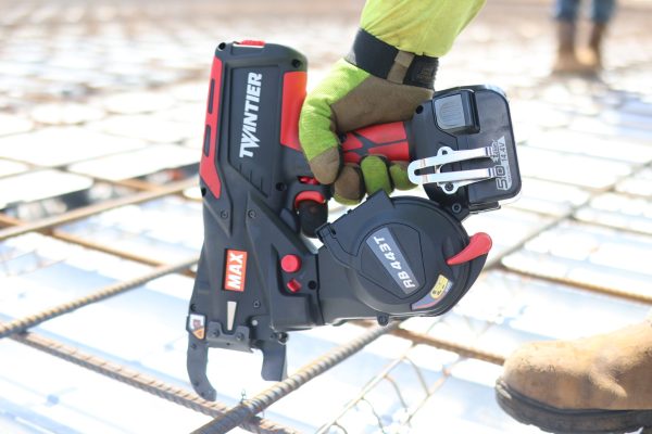 A worker using a Max TwinTier RB443T rebar tying tool to tie rebar on a commercial job site. The tool includes a battery and a belt hook.