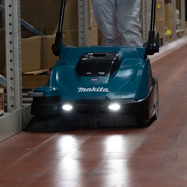 A worker is using the Makita VS001GZ 40V Cordless XGT Vacuum Sweeper to clean the floor in an industrial warehouse. A view of the front of the sweeper with headlights on.