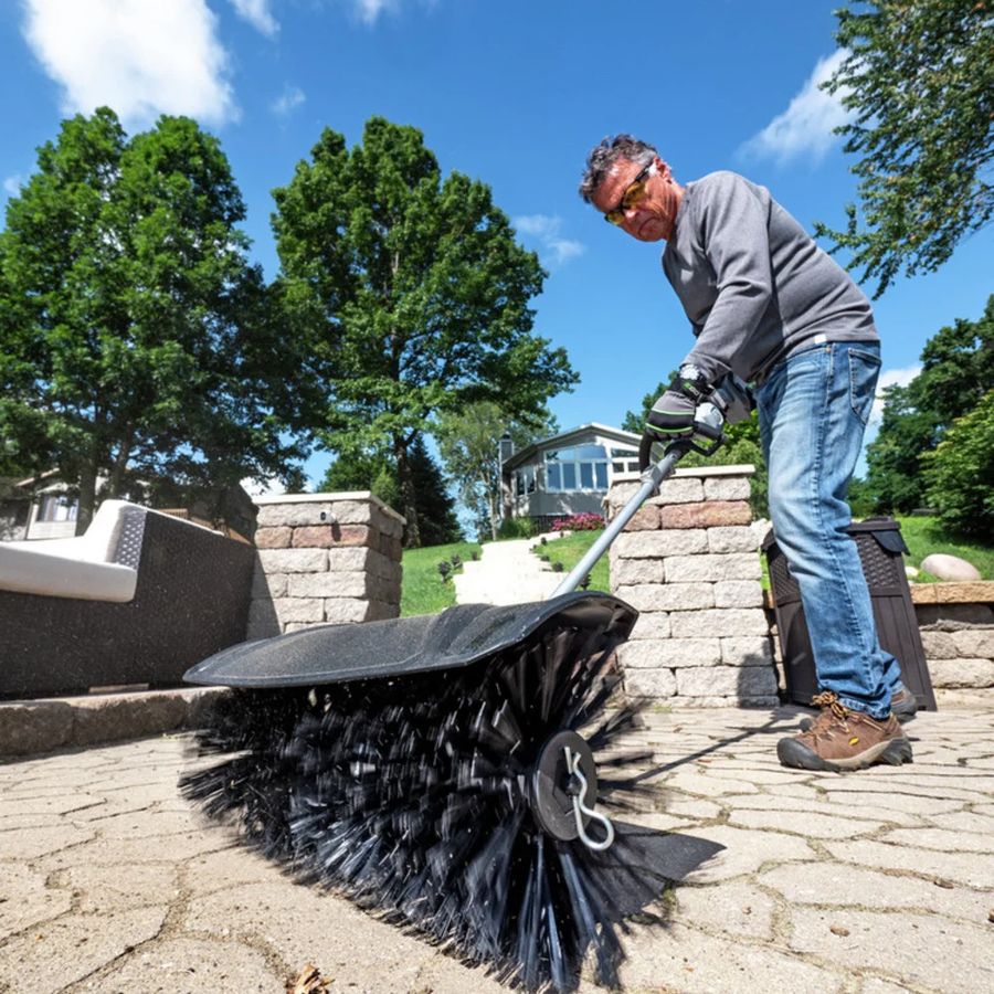 Person using the EGO BBA2100 bristle brush attachment on the EGO POWER+ Multi-Head System to sweep leaves and debris from a paved patio surface in a residential outdoor setting.