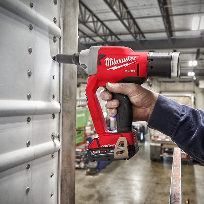 Milwaukee M18 FUEL 1/4″ blind rivet tool with ONE-KEY installing rivets into a corrugated metal panel, showing a worker’s hand operating the cordless rivet gun with M18 REDLITHIUM battery in an industrial workshop setting.