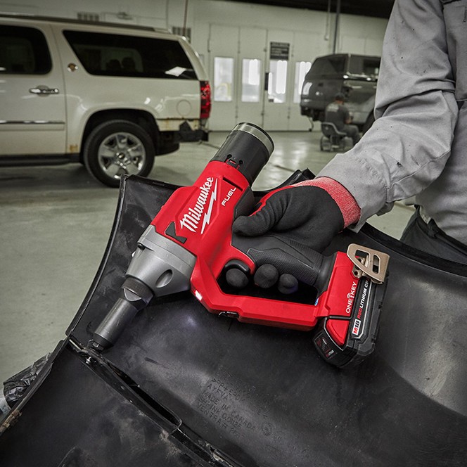 Milwaukee M18 FUEL 1/4″ blind rivet tool with ONE-KEY fastening a rivet into an automotive body panel, showing a gloved technician using the cordless rivet gun with M18 REDLITHIUM battery inside a vehicle repair shop.