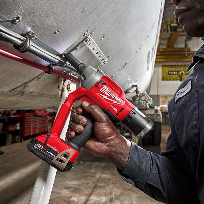 Milwaukee M18 FUEL 1/4″ blind rivet tool with ONE-KEY being used to install rivets on an aluminum aircraft-style panel, showing a gloved worker holding the cordless rivet gun with mandrel collector attached in an industrial shop environment.