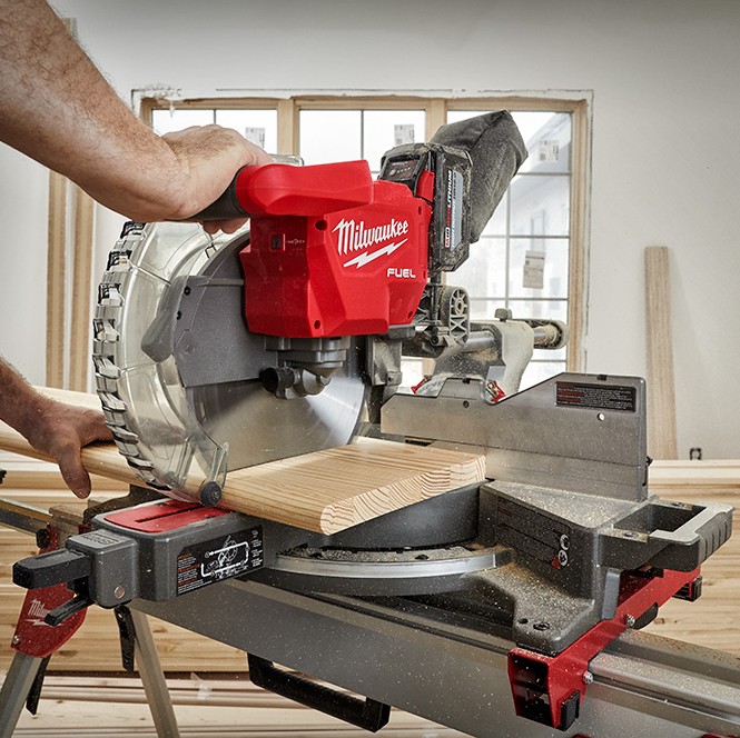 Milwaukee M18 FUEL 12-inch dual bevel sliding compound mitre saw cutting dimensional lumber indoors, shown in use by an operator with hands on the handle and workpiece, blade guard lowered, sliding rails engaged, and dust collection bag attached