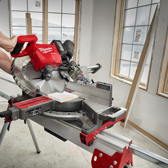 Milwaukee M18 FUEL 12-inch dual bevel sliding compound mitre saw in use indoors, shown making a crosscut on dimensional lumber with the saw head lowered, sliding rails extended, blade guard engaged, and dust collection bag attached on a portable stand