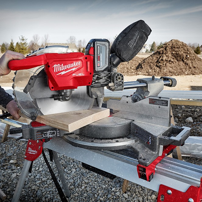 Milwaukee M18 FUEL 12-inch dual bevel sliding compound mitre saw cutting a board outdoors on a portable stand, shown mid-cut with the sliding rails extended, dust bag attached, and an M18 battery installed.