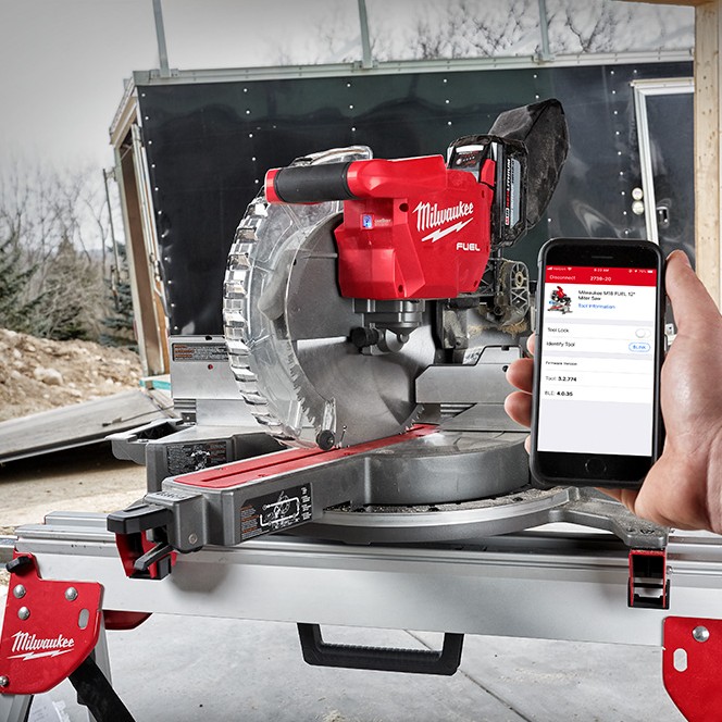 Milwaukee M18 FUEL 12-inch dual bevel sliding compound mitre saw on a jobsite stand, shown idle with blade raised while a smartphone displays the ONE-KEY app used to track and manage the saw.