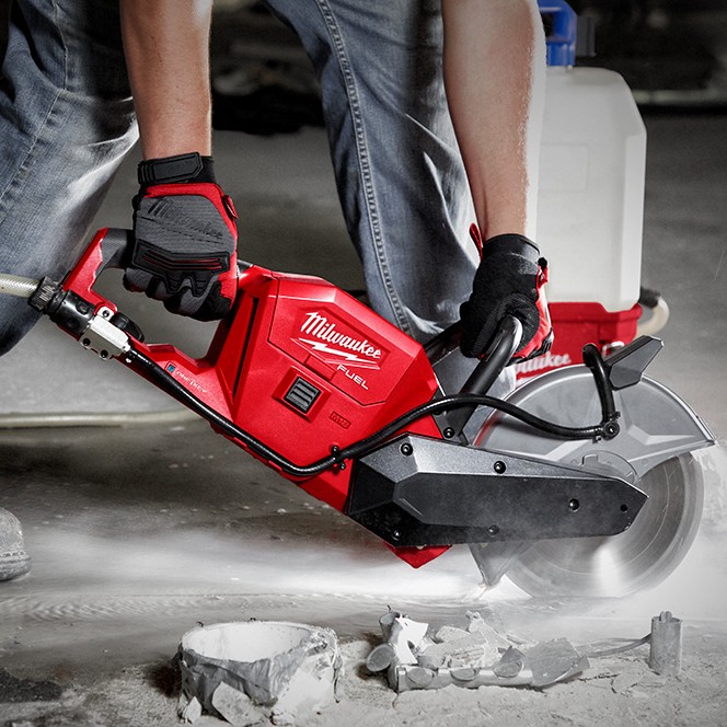 Milwaukee M18 FUEL 9-inch cordless cut-off saw cutting concrete indoors with water suppression, shown in use by a worker wearing gloves, with diamond blade engaged, visible water spray, and SWITCH TANK water supply connected for dust control