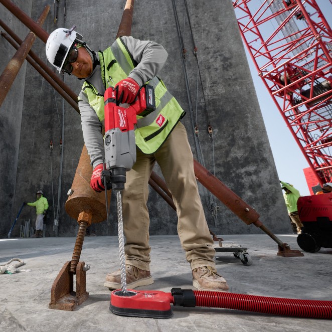 Worker wearing safety helmet, gloves, and high-visibility vest using a Milwaukee M18 FUEL SDS Plus D-handle rotary hammer with dust extraction shroud to drill into a concrete floor on an active construction site with steel supports and crane structure visible.