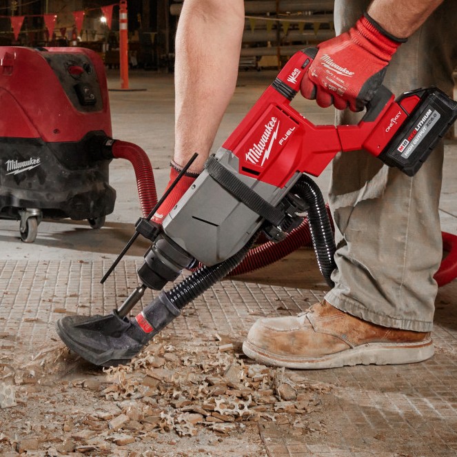 Worker using a Milwaukee M18 FUEL SDS Plus D-handle rotary hammer with attached dust extraction shroud to drill into a concrete floor indoors, with debris visible and a vacuum system connected for dust control.