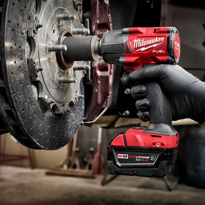 Milwaukee M18 FUEL 3/8 in. mid-torque impact wrench removing nut from a vehicle brake rotor, shown in use with an M18 REDLITHIUM XC5.0 battery in an automotive shop setting.