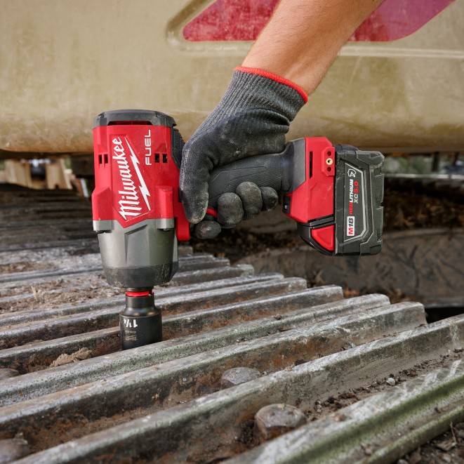 Milwaukee M18 FUEL 1/2 in. high-torque impact wrench loosening or tightening a bolt on a tracked equipment pad, shown in use with an M18 REDLITHIUM XC battery on heavy machinery undercarriage.