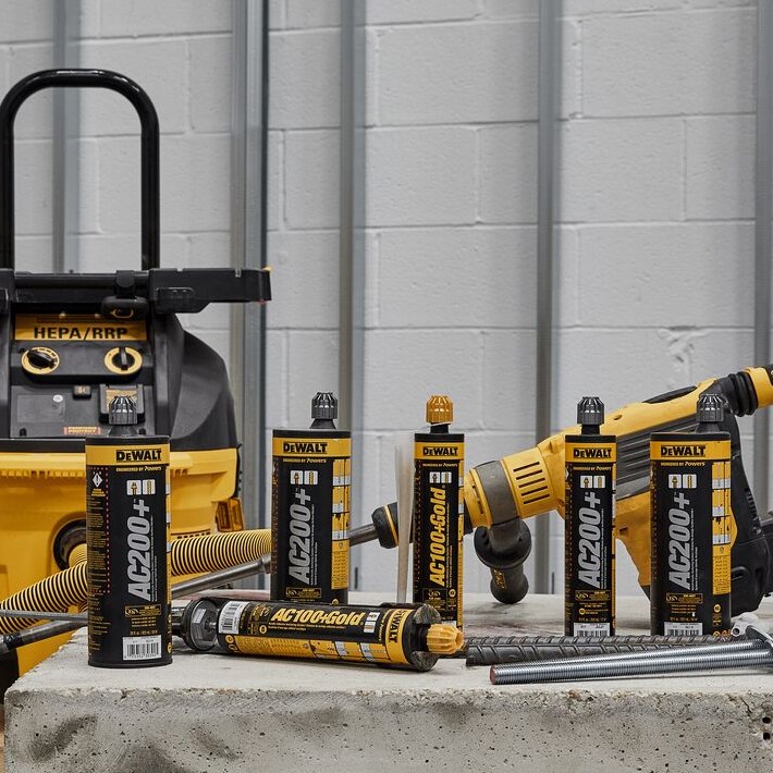 Multiple DEWALT AC200+ and AC100+ Gold anchoring adhesive cartridges displayed on a concrete slab alongside a rotary hammer drill, HEPA dust extractor, threaded rod, and rebar, illustrating a professional concrete anchoring jobsite setup.