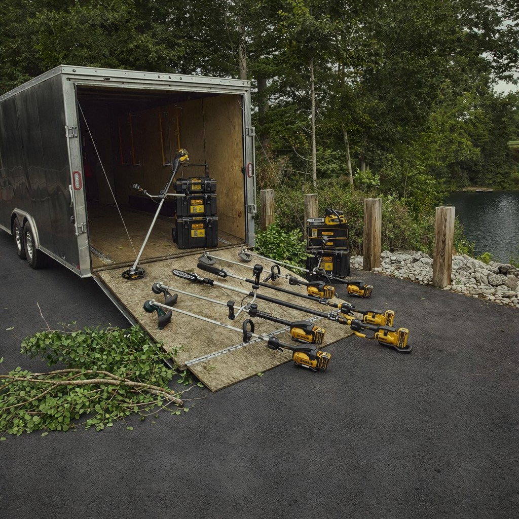 DEWALT cordless landscaping tools and attachment system laid out on a trailer ramp, including multiple trimmers and power heads, with storage cases and a wooded lakeside background.