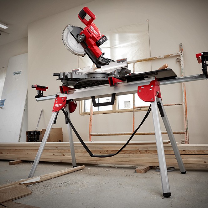 Milwaukee M18 FUEL 7-1/4 in dual bevel sliding compound mitre saw mounted on folding stand in interior jobsite with stacked lumber, ladder and unfinished drywall walls visible