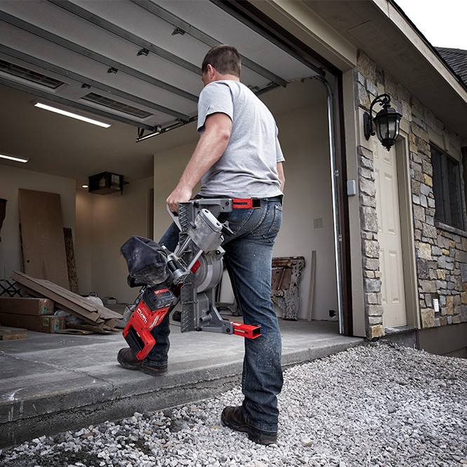 Worker carrying Milwaukee M18 FUEL dual bevel sliding compound mitre saw up step into open garage, saw held by top handle with dust bag attached, residential jobsite background with stone exterior and building materials inside garage