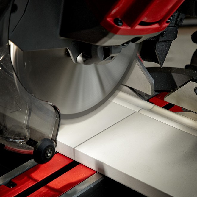 Close-up of Milwaukee M18 FUEL 7-1/4 in dual bevel sliding compound mitre saw blade cutting white trim board on red saw table, blade guard and fence visible during precision cut