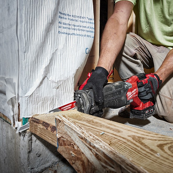 Milwaukee M18 FUEL Sawzall reciprocating saw with XC6.0 battery cutting pressure-treated wood framing beside exterior wall sheathing and house wrap while operated by a gloved worker at a construction site