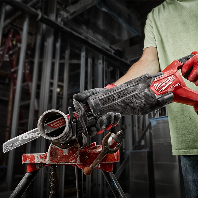 Milwaukee M18 FUEL Sawzall reciprocating saw cutting metal pipe secured in a pipe vise inside a commercial construction setting with exposed steel framing and a gloved operator holding the tool