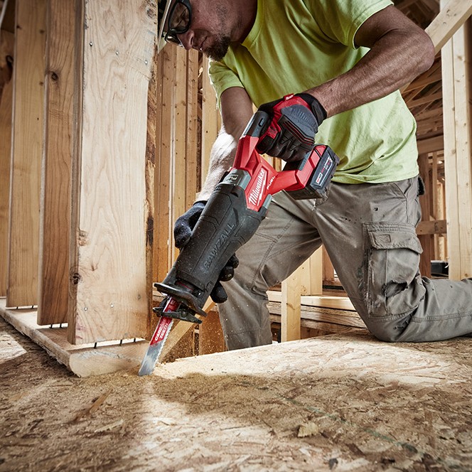 Milwaukee M18 FUEL Sawzall reciprocating saw with XC6.0 battery cutting wood subfloor inside a framed construction structure while held by a worker wearing gloves and safety glasses