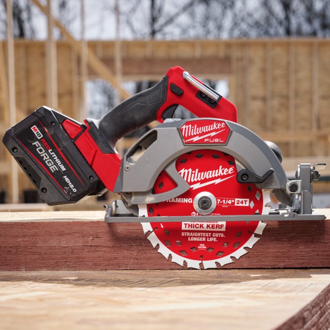 Milwaukee M18 FUEL 7-1/4 inch circular saw side view resting on dimensional lumber with FORGE HD12.0 battery installed, blade exposed, framing jobsite structure in background
