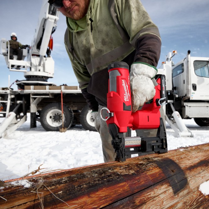 Utility worker driving fencing staples into a wooden utility pole using a Milwaukee M18 FUEL utility fencing stapler, with crane truck and snow-covered ground visible at a jobsite.