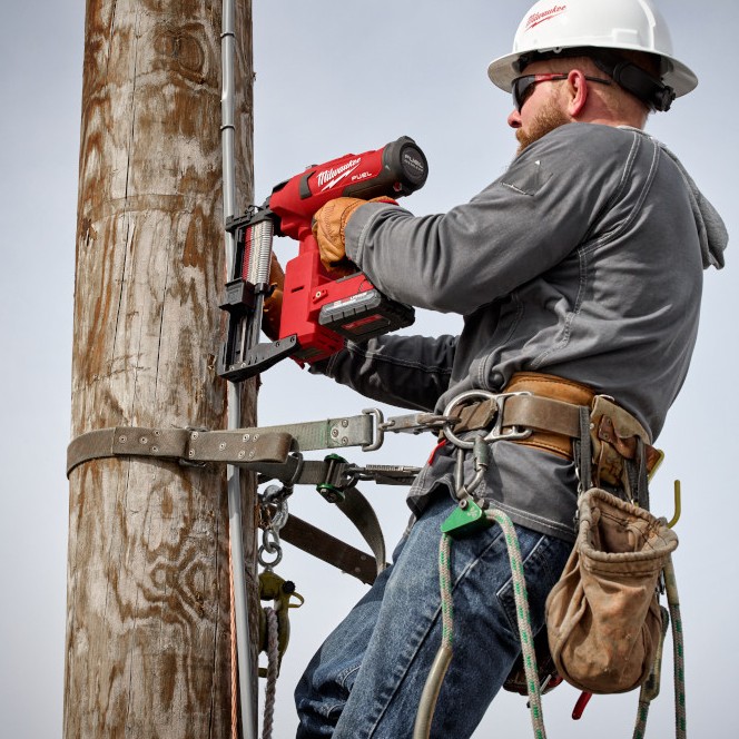 Utility lineman secured to a wooden utility pole using a safety harness while driving fencing staples with a Milwaukee M18 FUEL utility fencing stapler.