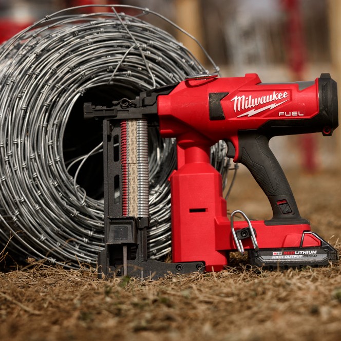 Milwaukee M18 FUEL utility fencing stapler with battery installed, standing on ground beside a large coil of galvanized fencing wire, with staple magazine visible and tool positioned upright outdoors.