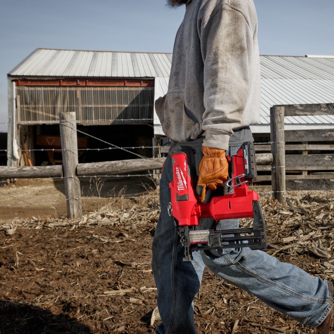 Worker carrying a Milwaukee M18 FUEL utility fencing stapler at a farm site, with fencing posts and a barn in the background, showing the tool held at the side while walking between fence lines.