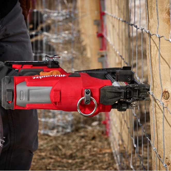 Close-up of a Milwaukee M18 FUEL utility fencing stapler driving a galvanized fence staple into a wooden fence post, with welded wire fencing held tight against the post during installation.