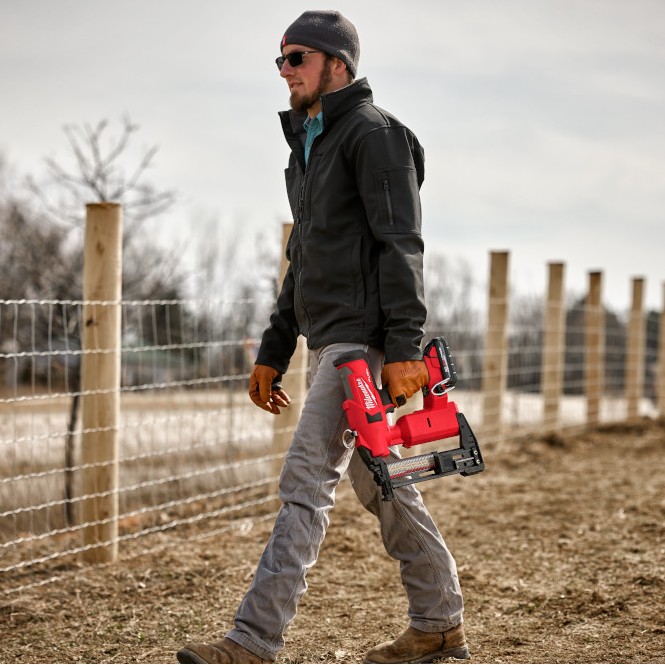 Worker walking along a fenced property line carrying a Milwaukee M18 FUEL utility fencing stapler, with installed wire fencing and wooden fence posts visible in an outdoor agricultural setting.