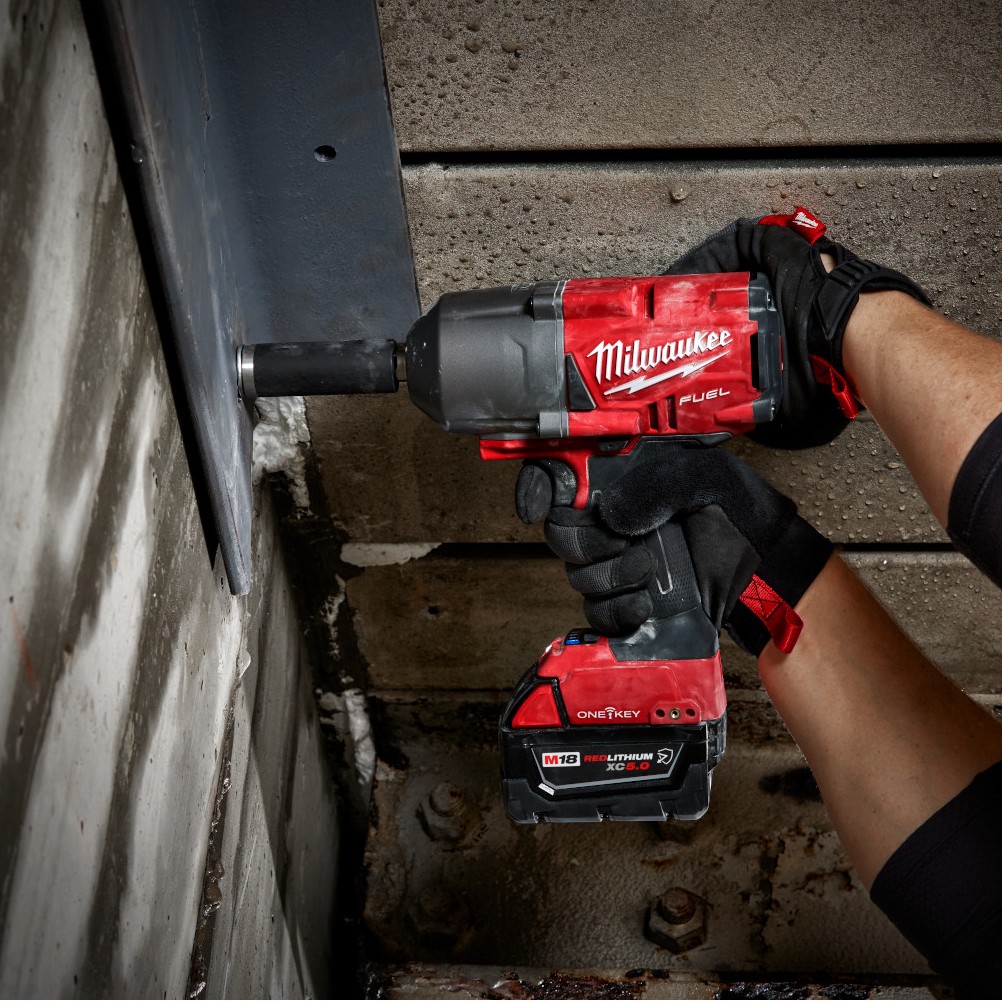Milwaukee M18 FUEL high-torque impact wrench with ONE-KEY and XC5.0 battery being used to tighten or loosen a large socket on a steel beam, held by gloved hands in an industrial concrete setting.