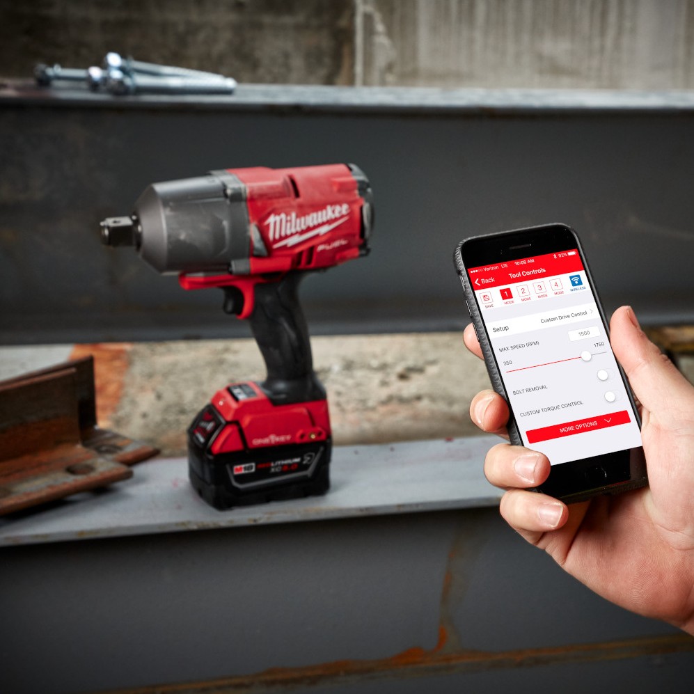 Milwaukee M18 FUEL high-torque impact wrench with battery resting on a steel beam, with a hand holding a smartphone displaying the ONE-KEY app interface beside the tool in an industrial setting.