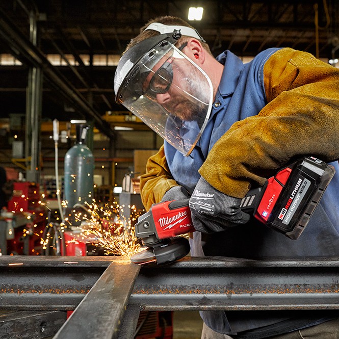 Milwaukee M18 FUEL 4-1/2 inch / 5 inch Braking Grinder with XC6.0 battery grinding steel beam in industrial workshop, operator wearing face shield, safety glasses, gloves, and protective sleeves with sparks visible during metal grinding.