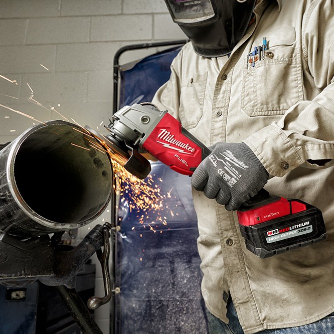 Milwaukee M18 FUEL cordless angle grinder with XC6.0 battery grinding a large steel pipe, producing sparks, held by a worker wearing gloves and a welding helmet in an indoor workshop setting