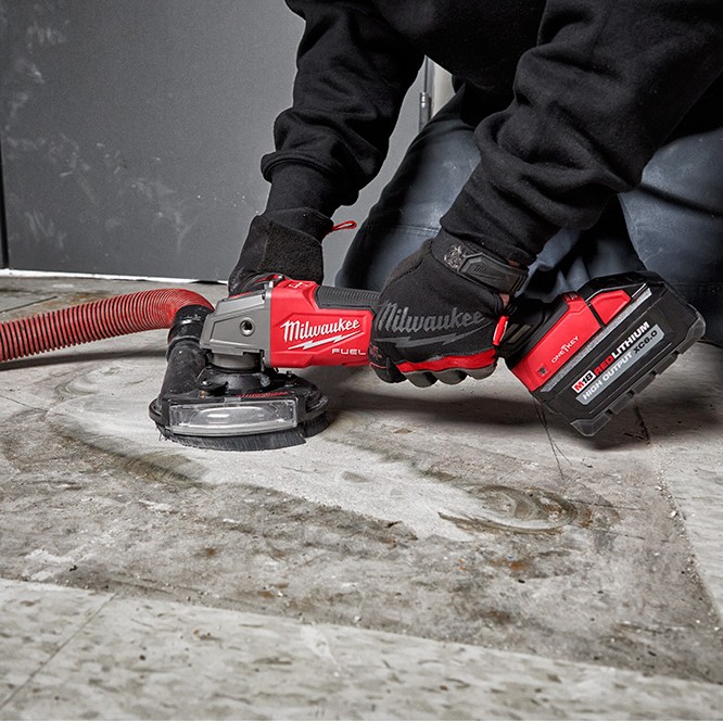 Milwaukee M18 FUEL 4-1/2 in grinder with dust shroud and vacuum hose attached, grinding concrete floor while operator kneels and holds paddle switch, M18 REDLITHIUM HIGH OUTPUT XC6.0 battery installed
