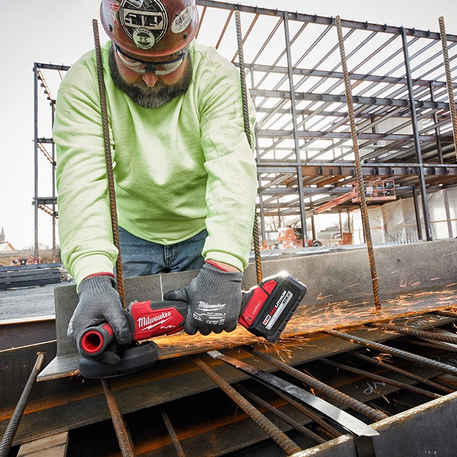 Milwaukee M18 FUEL cordless angle grinder with XC6.0 battery cutting steel rebar on a construction site, producing sparks, operated by a worker wearing gloves, safety glasses, and a hard hat in front of a structural steel frame building