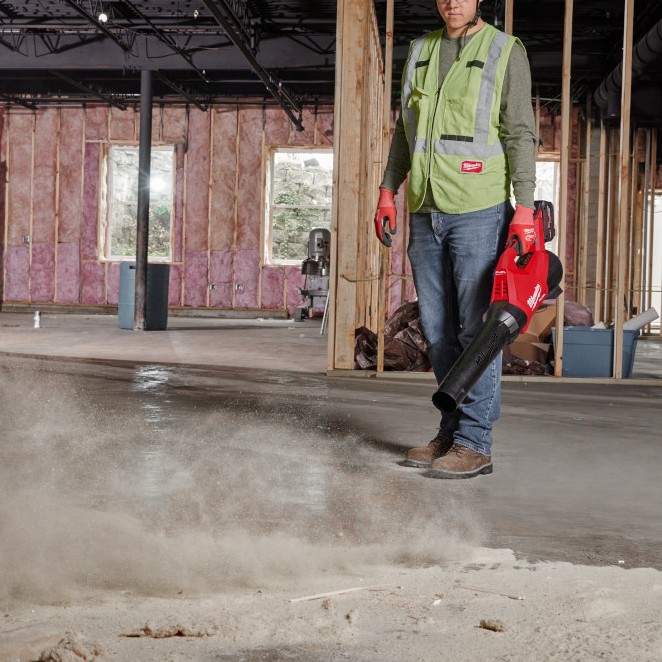 Milwaukee M18 FUEL cordless blower clearing dust from concrete floor inside unfinished construction interior with exposed framing and insulation