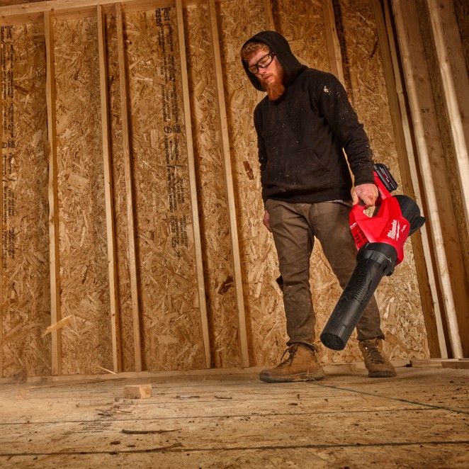 Milwaukee M18 FUEL cordless blower clearing sawdust and debris from plywood subfloor inside framed construction space with exposed studs