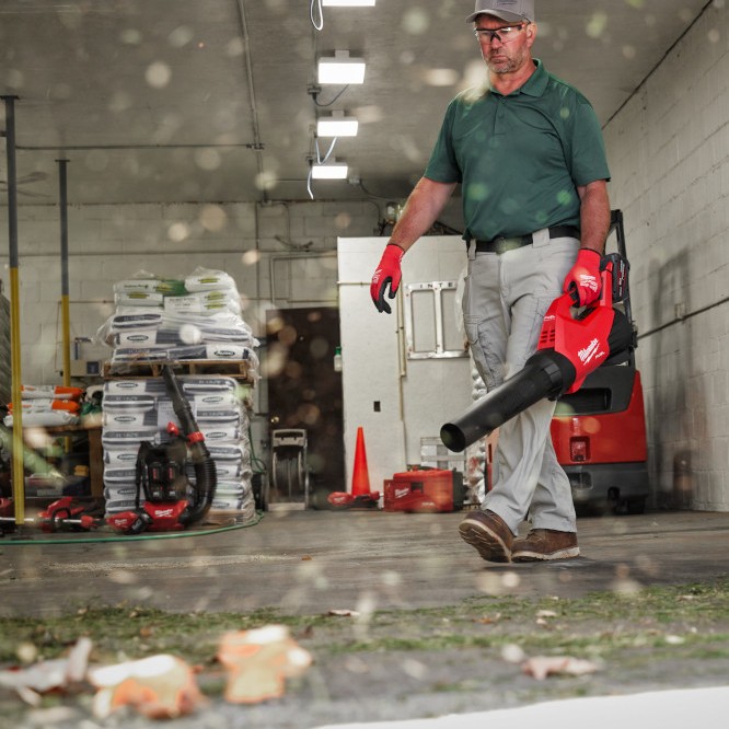Milwaukee M18 FUEL cordless blower clearing debris inside warehouse bay with worker walking past palletized materials and equipment