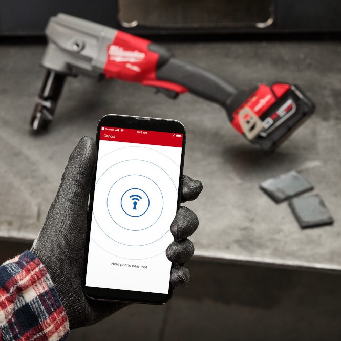 Gloved hand holding a smartphone showing Milwaukee ONE-KEY pairing screen, with a Milwaukee M18 FUEL nibbler resting on a metal workbench in the background.