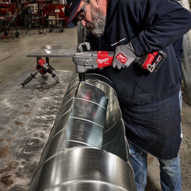 Tradesperson using a Milwaukee M18 FUEL nibbler to cut curved sections in large galvanized HVAC ducting inside a metal fabrication shop.