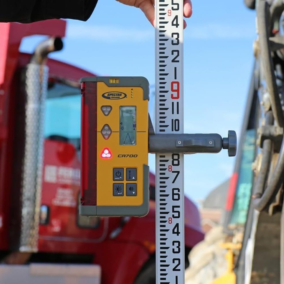 Spectra Precision CR700 laser receiver mounted to a grade rod with the clamp, showing digital readout and LED arrows, with a work truck and open sky visible in the background during outdoor grading.