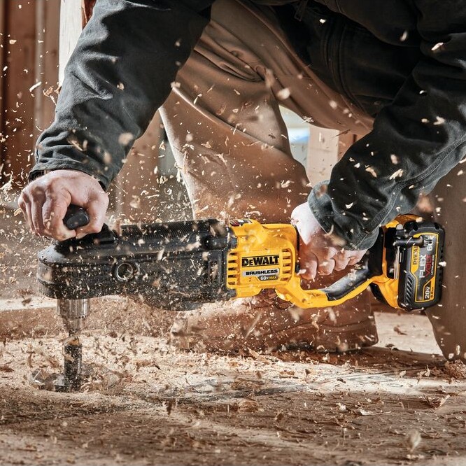 DEWALT FLEXVOLT stud and joist drill boring into wood floor with auger bit, operator kneeling with wood chips flying across construction site floor, cordless battery attached to rear handle