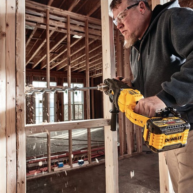 DEWALT FLEXVOLT in-line stud and joist drill boring large hole through wood wall framing with long auger bit, operator wearing safety glasses inside unfinished house framing with wood shavings flying