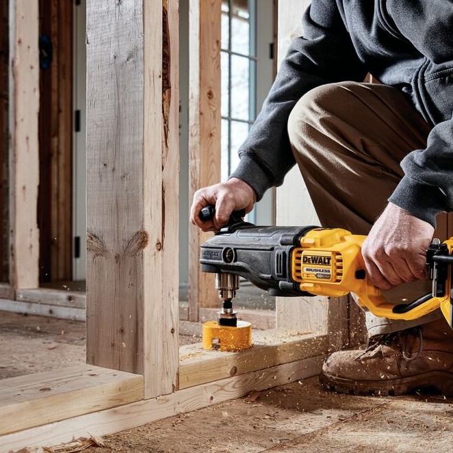 DEWALT FLEXVOLT in-line stud and joist drill with hole saw cutting through wood bottom plate, operator kneeling on construction floor inside unfinished framed structure with wood debris visible