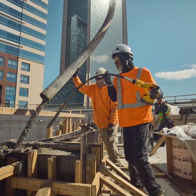 Two construction workers wearing hard hats and high-visibility shirts using a yellow DEWALT cordless concrete pencil vibrator to consolidate concrete flowing from a pump hose into wood formwork at an urban jobsite with high-rise buildings in background