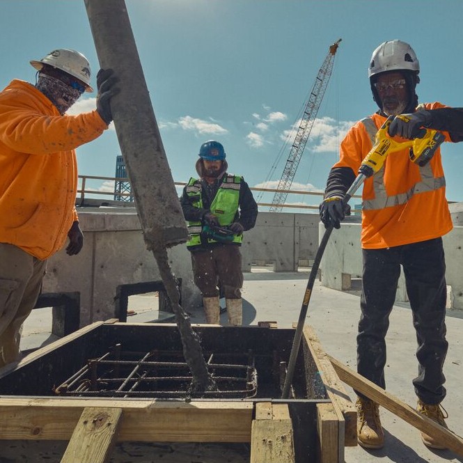 Construction workers pouring concrete into wood formwork while a worker uses a yellow DEWALT cordless concrete pencil vibrator to consolidate concrete around rebar, crane and jobsite structures visible in background