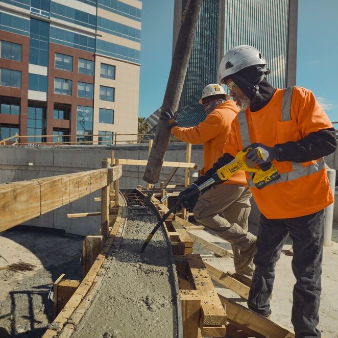 Two construction workers in hard hats and high-visibility shirts using a yellow DEWALT cordless concrete pencil vibrator to consolidate wet concrete inside curved wood formwork while concrete pours from a pump hose, city buildings visible in background