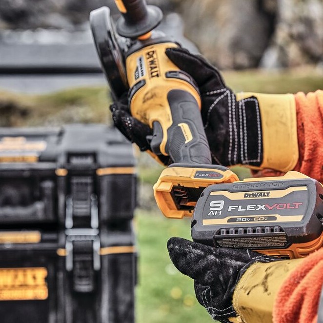 DEWALT 60V MAX brushless angle grinder with FLEXVOLT 9Ah battery being inserted, close-up of battery connection and rear handle, toolboxes blurred in background.