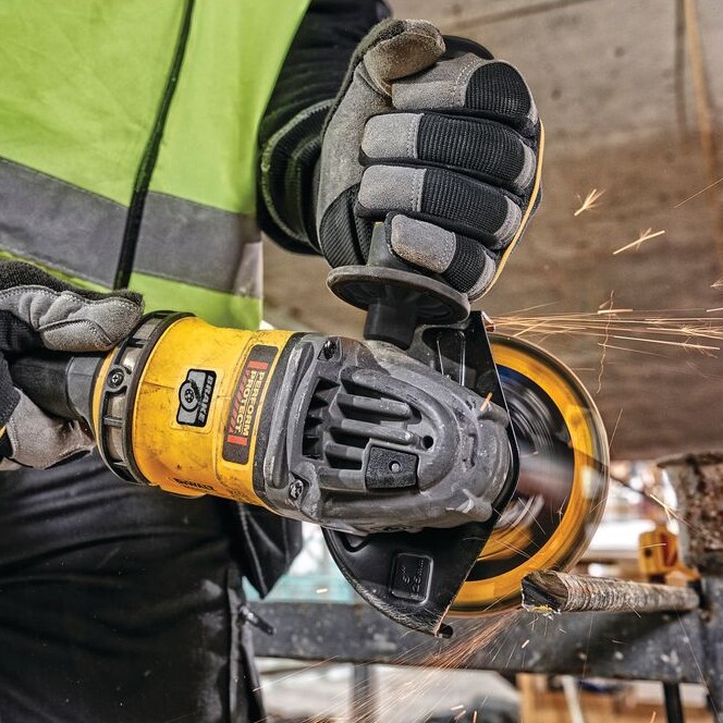 Worker using DEWALT cordless angle grinder with side handle to cut rebar, sparks visible, wearing gloves and high-visibility vest on a construction site.